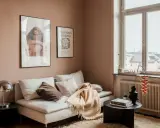 Un divano bianco accogliente con cuscini e una coperta color **rosa pastello** su un tappeto soffice. Alle pareti, due quadri e una finestra con vista.