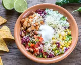 Bunter Taco Salat mit Hähnchen, Reis, Bohnen, Mais, Tomaten, Käse und Sauerrahm. Tortilla-Chips und Limetten daneben.