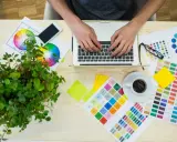 Grafik pracuje przy laptopie, otoczony próbkami kolorów i kołem barw. To idealne drukarskie tło dla kreatywnych projektów.