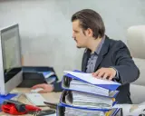Mężczyzna w garniturze pracuje przy biurku, przeglądając dokumenty. Przed nim stos segregatorów, komputer i czerwony telefon. To obraz codziennego postępowania administracyjnego.