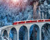 Un treno rosso attraversa un viadotto innevato, parte del suggestivo treno Bernina percorso, tra alberi coperti di neve e rocce imponenti.