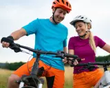Uśmiechnięta para w kaskach i sportowych strojach, gotowa na przejażdżkę rowerami elektrycznymi. Wygodne akcesoria do roweru elektrycznego zapewniają komfort.