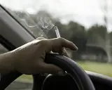 Mano sujetando un cigarrillo encendido sobre el volante. El humo se eleva mientras se puede fumar conduciendo.