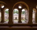Vue à travers les arches de l'abbaye du Thoronet, un cloître en pierre avec un jardin verdoyant et des bâtiments anciens.