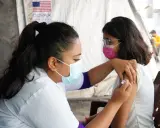 En sjuksköterska ger en vaccination i Nepal. Bakom syns en banderoll med texten COVAX och en amerikansk flagga.