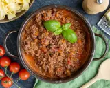 Herzhaftes Bolognese Rezept in einem Topf, garniert mit Basilikum. Daneben frische Tomaten, Bandnudeln und Parmesan.