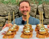 Hombre sonriente presenta cazuelitas de aperitivo con calamares y alioli sobre pan tostado, decoradas con brochetas.