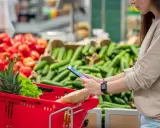 Donna al supermercato con carrello rosso, usa il telefono per i quantificatori spiegazione. Verdure sullo sfondo.