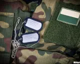 Dwa nieśmiertelniki na mundurze moro. Jeden z nich zawiera napis "MILITARIA.PL SHOOTING & OUTDOOR".