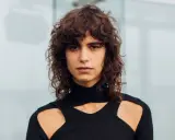 Ein Porträt einer Person mit voluminösem, welligem Haar und einem schwarzen Oberteil mit Cut-outs.
