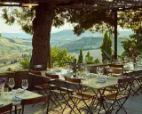 Tavoli apparecchiati sotto un pergolato con vista sulle dolci colline dell'entroterra toscana. Un invito a gustare la cucina locale.