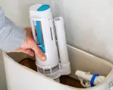 Mano instalando el mecanismo de descarga, una de las partes de una cisterna de baño. Se ve el flotador y la tubería de llenado.