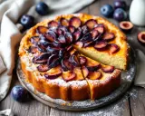 Gâteau aux prunes, saupoudré de sucre glace, avec des prunes fraîches disposées en rosace. Une part est coupée, révélant la texture moelleuse.