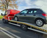Ciemnoszary samochód na lawecie, przygotowany do konfiskaty. Pomarańczowy pojazd pomocy drogowej stoi na poboczu drogi.
