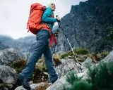 Kobieta z plecakiem wyrusza na wypad w góry, wspierając się kijkami trekkingowymi po kamienistym szlaku.