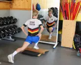 Ein Athlet im "Germany Triathlon"-Shirt macht Ausfallschritte im Athletik Training. Hanteln und Trainingsgeräte im Hintergrund.