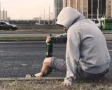 Mężczyzna w kapturze siedzi przy drodze z butelką piwa. Jego postawa sugeruje walkę z nałogiem, być może potrzebuje pomocy terapeuty od uzależnień.