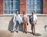 Three men in stylish suits stand outside a brick building, ready for a cocktail attire wedding.
