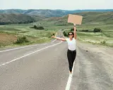 Kobieta z kartonem i kciukiem w górze na pustej drodze. Uczy się, jak łapać stopa, podróżując przez malownicze krajobrazy.