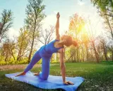 Kvinna utför yoga utomhus i en skogsmiljö. Solen skiner genom träden.