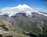 Majestatyczny Elbrus, najwyższy szczyt Europy, góruje nad skalistymi zboczami i lodowcami. W dole widać niewielką osadę, która pokazuje, gdzie leży Elbrus.