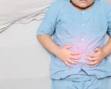 Niño con pijama azul, acostado en cama blanca, se agarra la barriga con las manos. Se visualiza un área rosada en su abdomen, indicando dolor barriga niños.