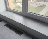 Jasny parapet z płytek wewnątrz, z widokiem na plac zabaw. Obok laptop i kratka wentylacyjna.