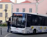 Ein Scott-Bus in Portugal hält an einer Haltestelle. Touristen steigen ein, während im Hintergrund bunte Gebäude und ein historischer Brunnen zu sehen sind.