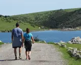 Ein Paar wandert auf einem Schotterweg am Ufer eines Sees. Grüne Hügel und Felsen prägen die **Spanien Natur**.