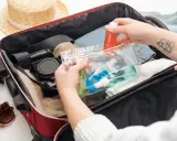 Ręce pakują kosmetyki w przezroczystej kosmetyczce do czerwonej walizki, obok słomkowego kapelusza i okularów.