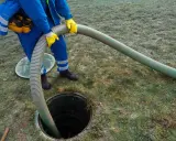 Pracownik w niebieskim kombinezonie i żółtych rękawicach wykonuje czyszczenie szamba, wkładając gruby wąż do otwartego włazu.