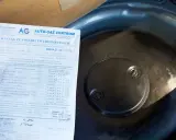 Fragment dokumentu z danymi homologacji butli LPG obok okrągłej pokrywy w bagażniku samochodu.