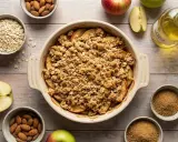 Crumble aux pommes dans un plat, entouré de pommes, d'amandes, de flocons d'avoine et de sucre brun.