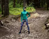 Kobieta w lesie, w kolorowych legginsach i zielonej bluzie, pokazuje, co daje stretching: elastyczność i siłę do pokonywania górskich ścieżek.