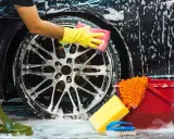 Persona con guantes amarillos detallando la llanta de un coche negro con espuma, mostrando cómo lavar el coche a mano. Cubo rojo y esponjas cerca.