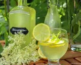 Erfrischendes Glas mit selbstgemachtem Holunderblütensirup, Zitrone und Minze. Daneben Flaschen mit dem Sirup und frische Holunderblüten.
