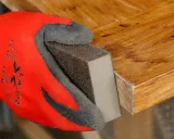 Mano con guante rojo lijando borde de puerta de madera para rebajarla.