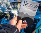 Dłoń trzyma książeczkę "Korona Gór Polski" na tle zimowego krajobrazu.