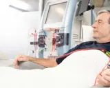 Starszy mężczyzna leży pod kocem, podłączony do aparatury do dializy. Jego ręka spoczywa na podłokietniku, a obok widać maszyny i czerwone wężyki.