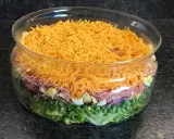 Une salade 7 étages colorée dans un grand bol en verre, avec des couches de laitue, petits pois, oignons rouges, jambon, œufs durs et fromage râpé.