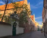Ulica z brukowaną nawierzchnią prowadzi do zabytkowego Collegium Maius. Złote światło słońca oświetla ceglane mury i pomarańczowe dachy.
