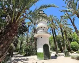 Torre blanca con balcón y puerta verde, rodeada de palmeras y vegetación exuberante. Un lugar encantador que ver en Elche.