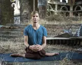 Man i blå tröja sitter i lotusställning på en yogamatta i ruiner. En stunds natthiko meditation i en övergiven miljö.