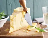 Ein Stück Parmesan, perfekt für vegetarische Gerichte, wird mit einem Käsemesser angeschnitten. Daneben ein Glas Weißwein und ein Salat.