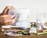 Ręce ozdabiające wazon techniką decoupage. Na stole nożyczki, pędzel, serwetki z lawendą i klej.