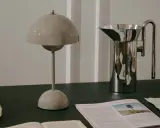 En beige lampa i studio lawahl-stil står bredvid en blank vattenkanna. En tidning med ett träd på omslaget ligger framför.