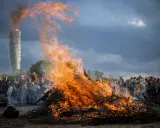 Stort bål med flammor som slår upp mot en dramatisk himmel. En folkmassa samlas för att bevittna ett traditionellt valborg firande.