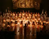 Stor grupp barn och ungdomar i vita kläder och luciakronor, med ljus i händerna, under ett lucia firande i Stockholm.