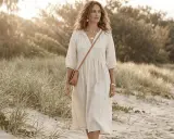 A woman walks on a beach in a flowing white dress, a tan crossbody bag, and gold jewelry. Her look embodies a natural, soft style, perfect for a David Kibbe test.