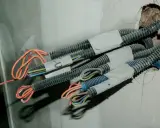 Przewody elektryczne w peszlach wychodzące ze ściany, gotowe do połączenia. Widać kolorowe żyły: pomarańczowe, niebieskie, brązowe, zielono-żółte.
